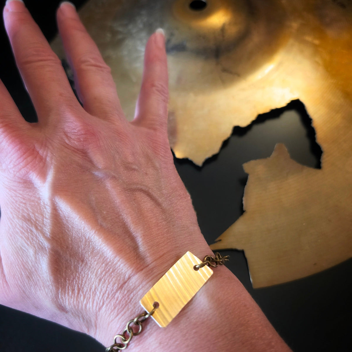 Partial view of a person's wrist wearing a gold-toned cymbal piece bracelet with chain links , against a dark background with a blurred brass cymbal.
