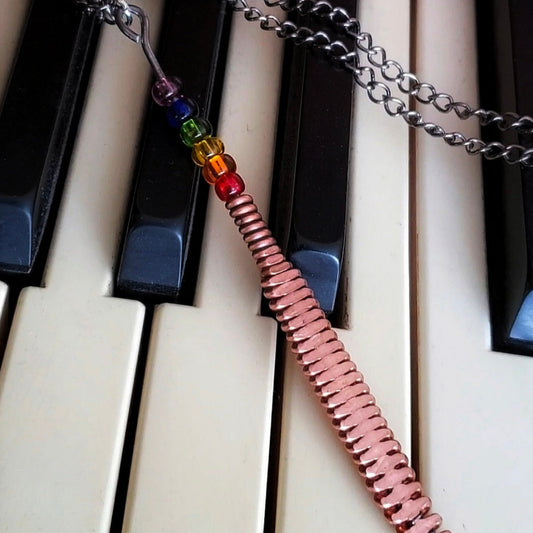 Hammered piano string pendant with 6 LGBTQ flag coloured beads with chain on a piano