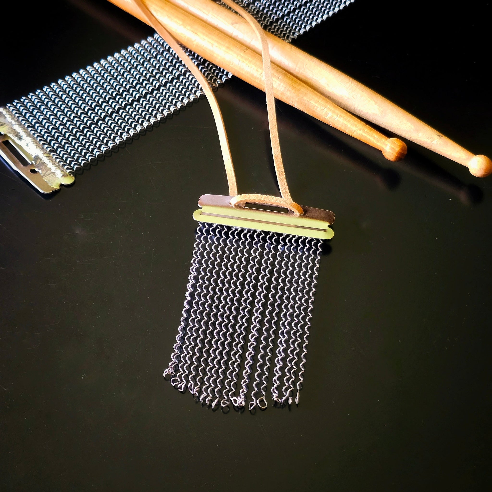 Closeup of a snare drum string fringe necklaces with light brown suede straps displayed on a black surface with wooden drumsticks and snare drum strings the background.