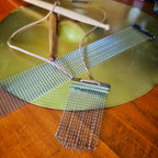 Snare drum string fringe necklace with light brown suede straps displayed on a cymbal with wooden drumsticks and snare drum strings the background.