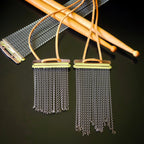 Two handmade snare drum string fringe necklaces with light brown suede straps (one longer than the other), displayed on a black surface with wooden drumsticks and snare drum strings the background.