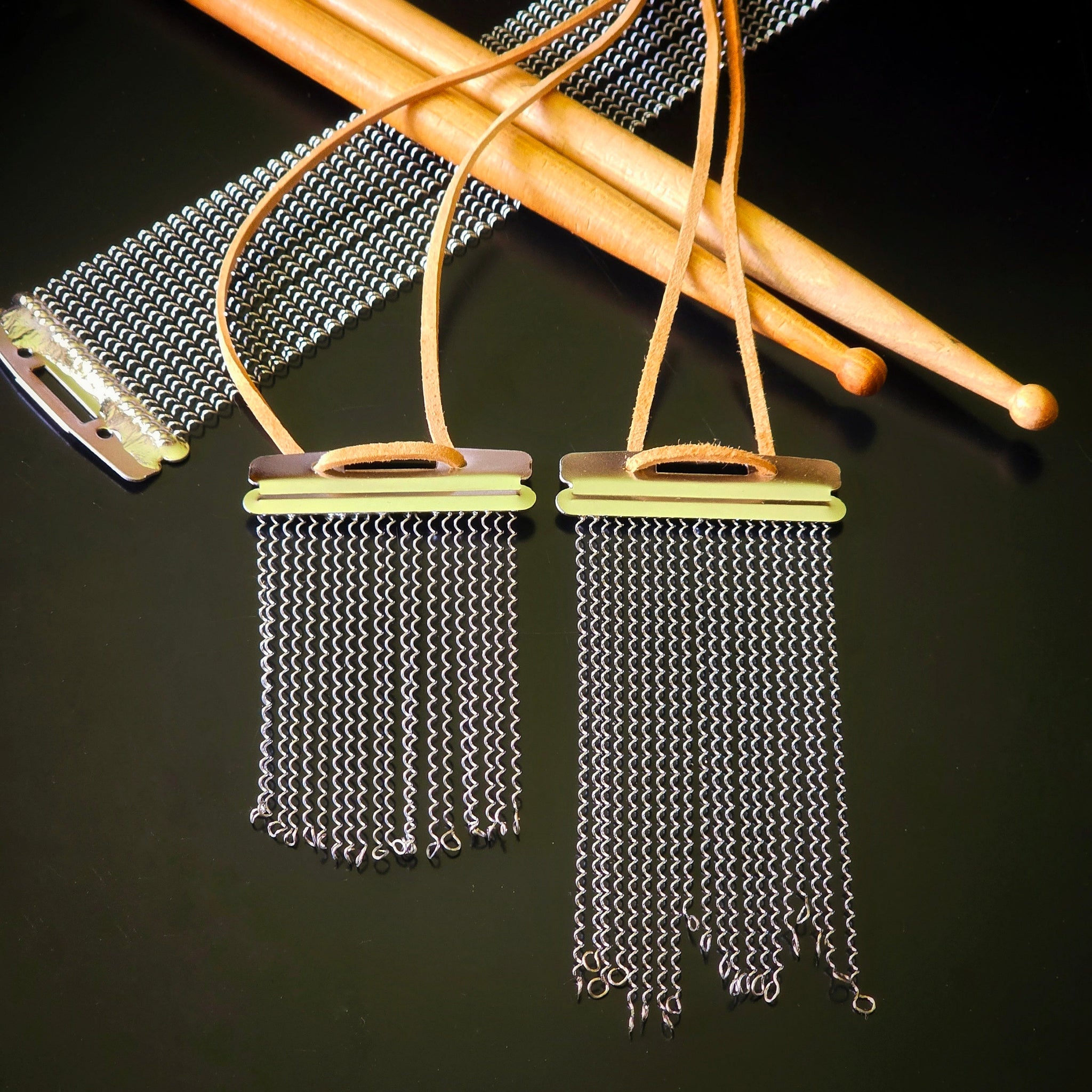 Two handmade snare drum string fringe necklaces with light brown suede straps (one longer than the other), displayed on a black surface with wooden drumsticks and snare drum strings the background.