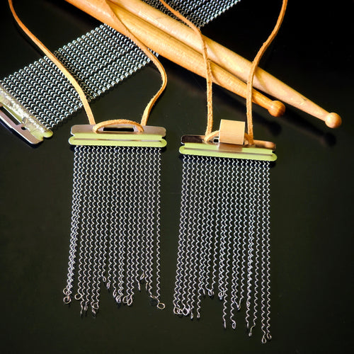Two handmade snare drum string fringe necklaces with light brown suede straps (one with and one without a drumstick bead), displayed on a black surface with wooden drumsticks and snare drum strings the background.