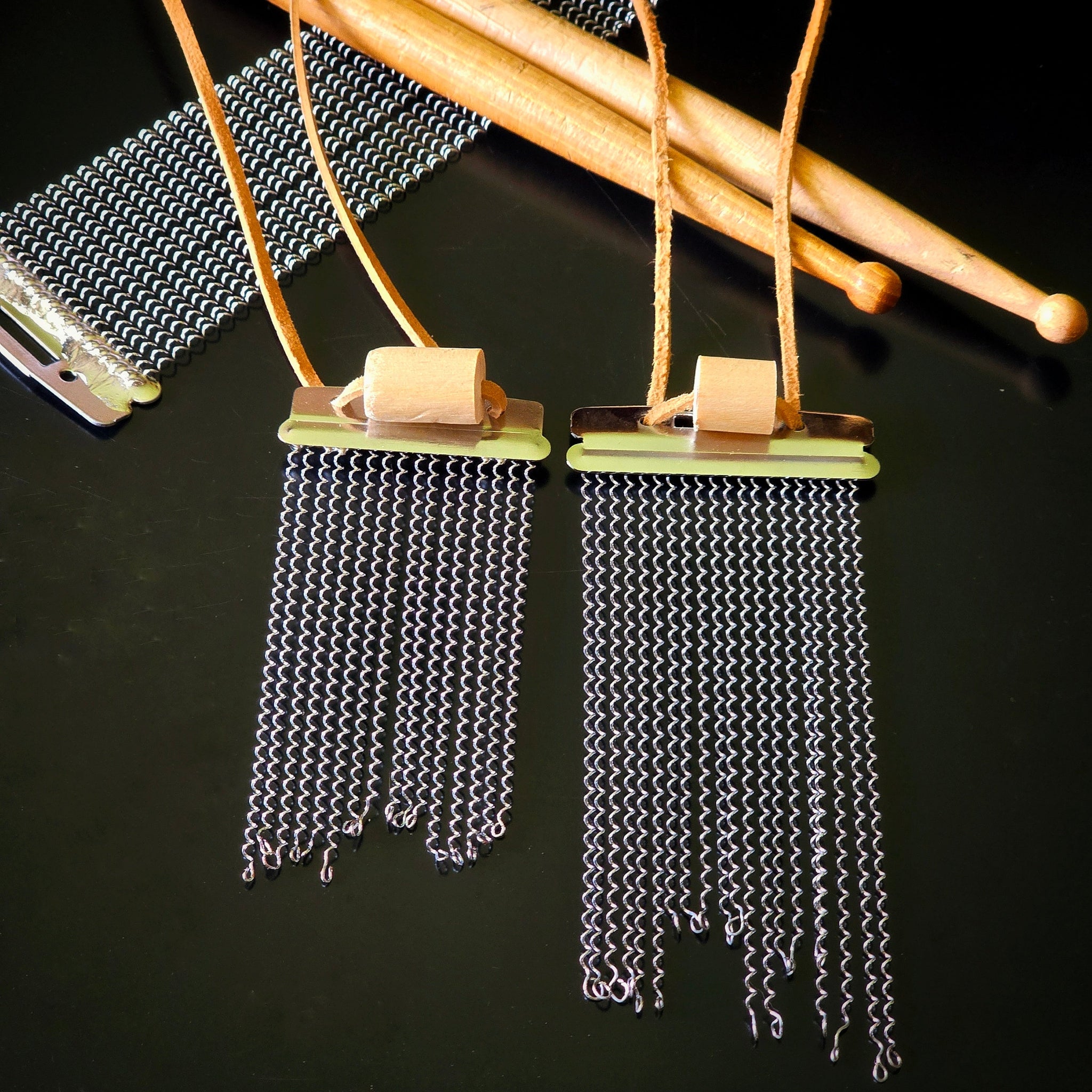 Two handmade snare drum string fringe necklaces (one shorter than the other) with light brown suede straps (both with a drumstick bead), displayed on a black surface with wooden drumsticks and snare drum strings the background.