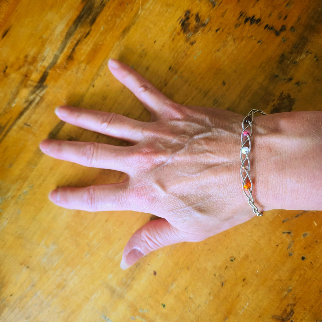 Woman's hand and wrist wearing a guitar string bracelet with 5 beads representing the colours of the lesbian pride flag 