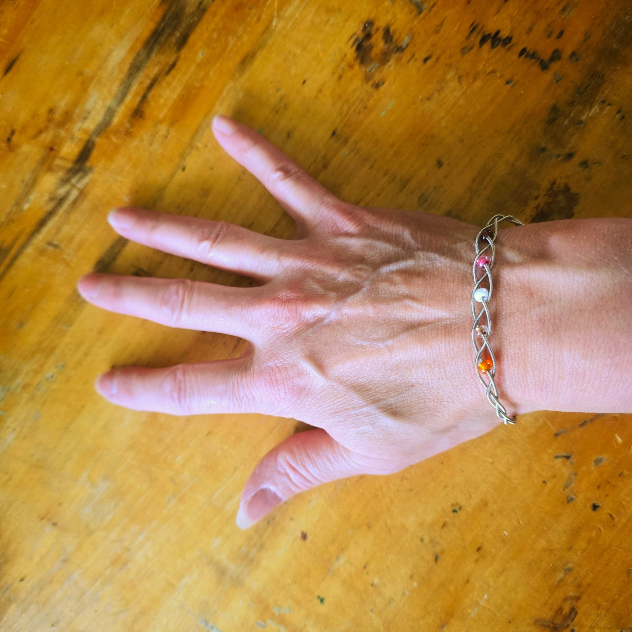 Woman's hand and wrist wearing a guitar string bracelet with 5 beads representing the colours of the lesbian pride flag 