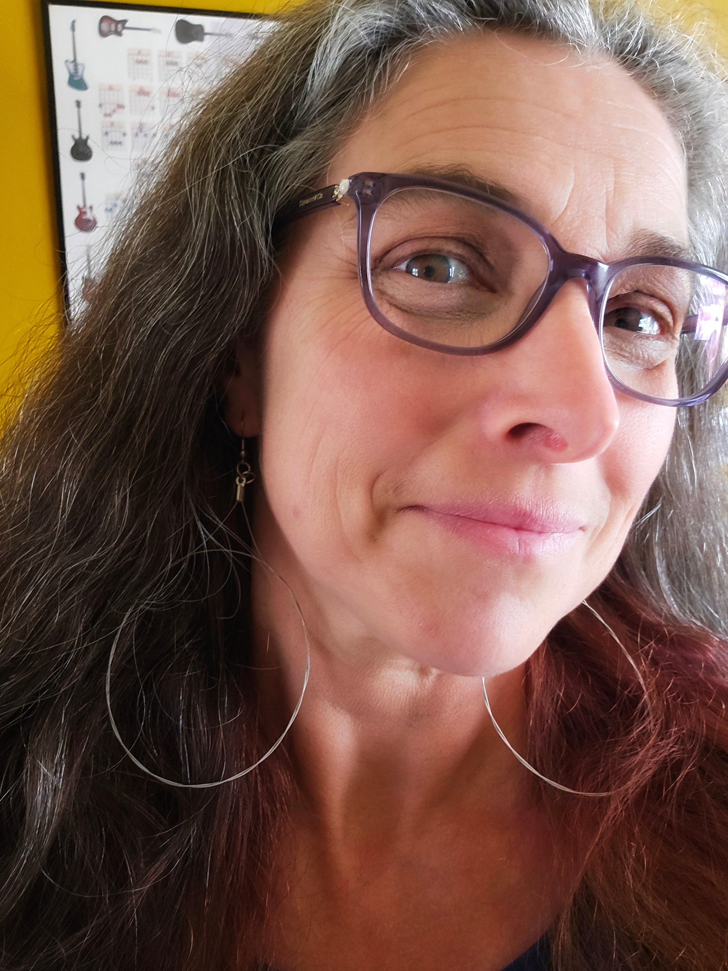 Woman wearing glasses  and large guitar string earrings with a yellow wall and guitar poster in the background