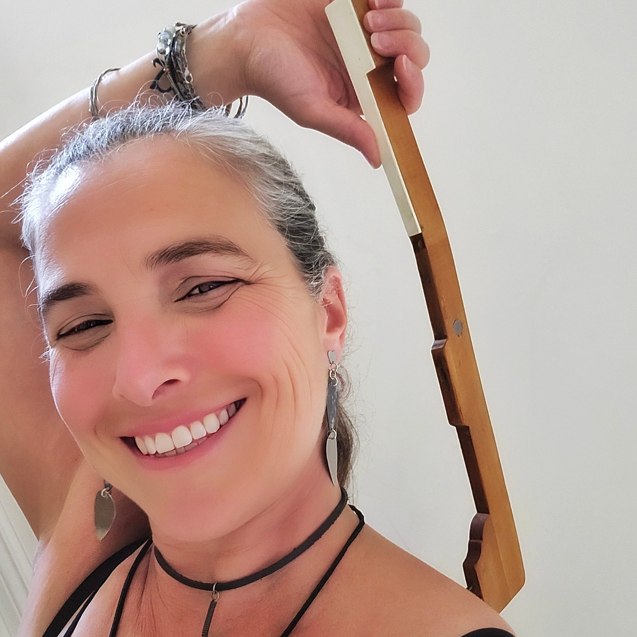 Woman with gray hair smiling, wearing a black top and earrings using an upcycled piano key backscracher