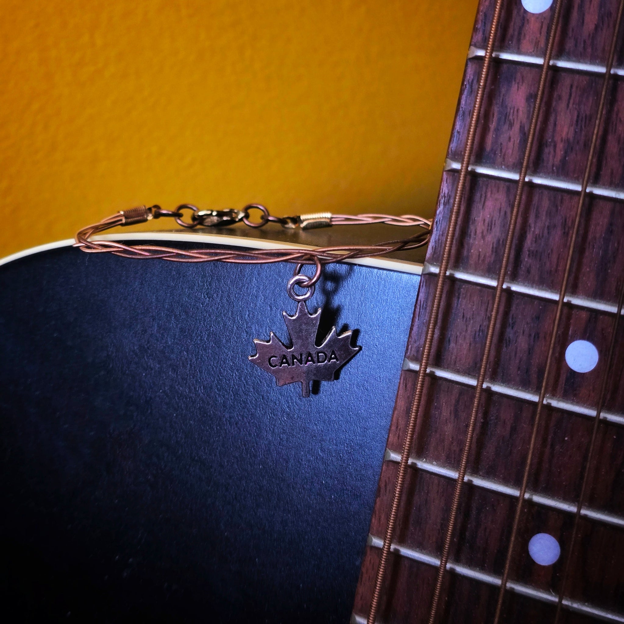 bronze coloured guitar string bracelet with canada maple leaf pendant on black guitar with yellow background