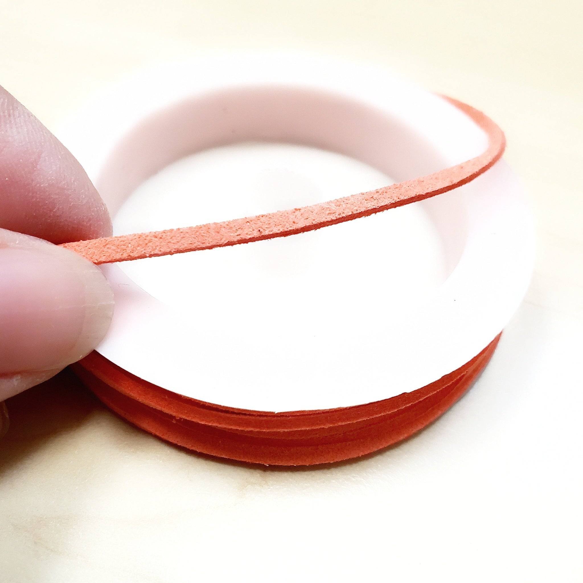 fingers holding a string of salmon coloured faux-suede coming from a white bobbin