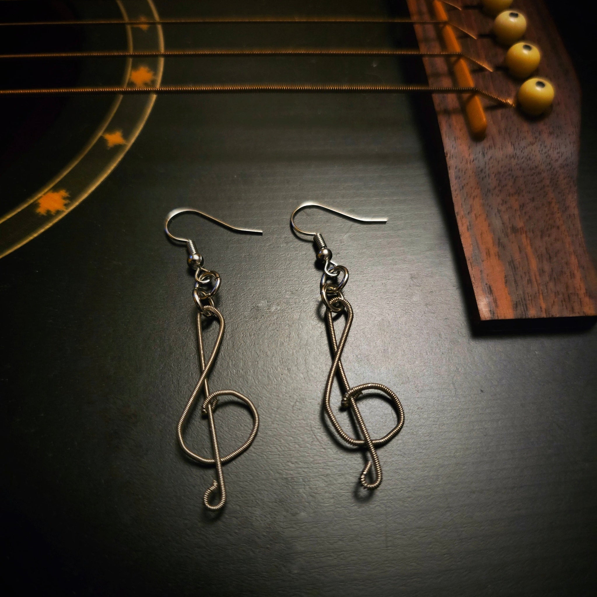 pair of earrings in the shape of treble clefs, made from upcycled guitar strings lying on the body of a black guitar