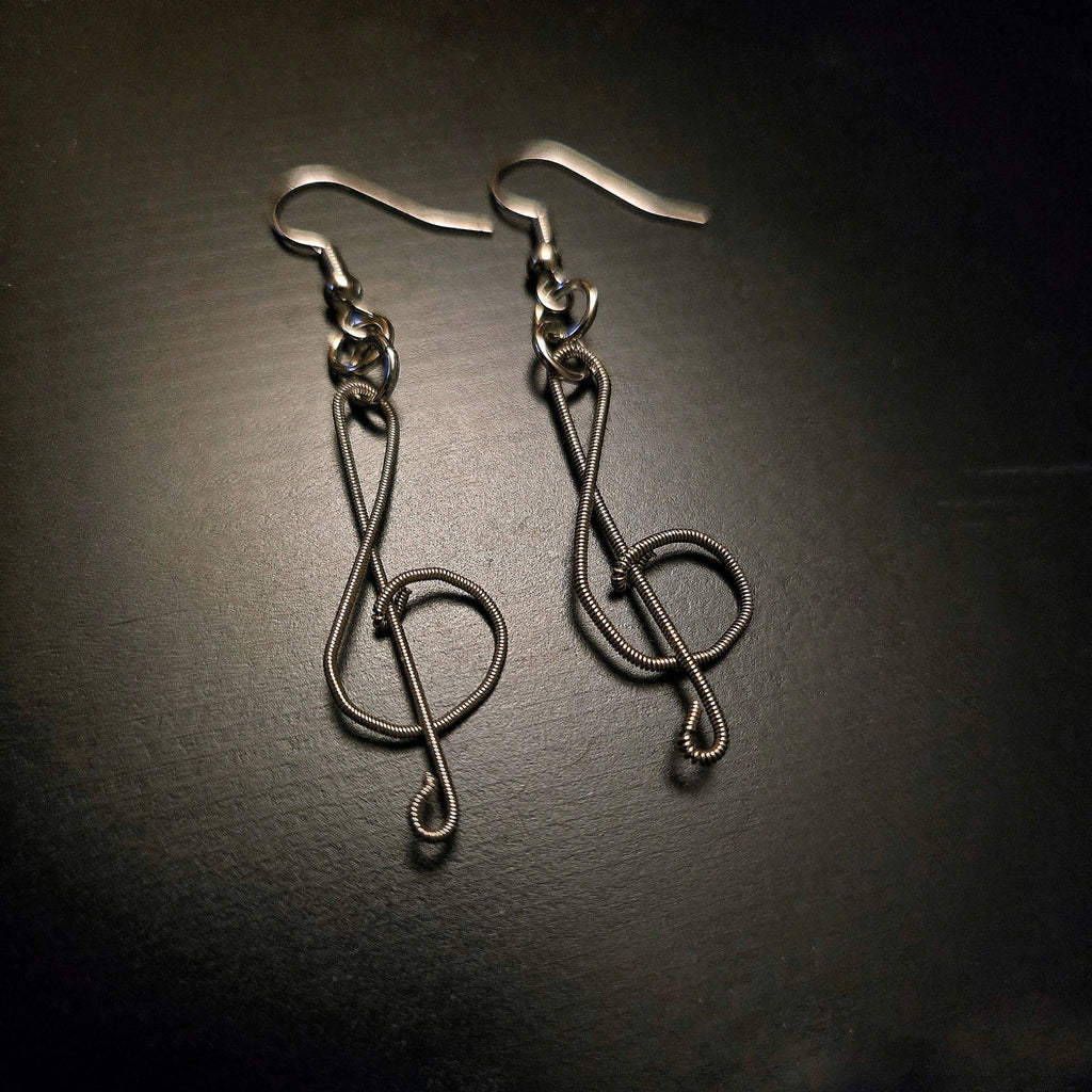 pair of earrings in the shape of treble clefs, made from upcycled guitar strings lying a black background 