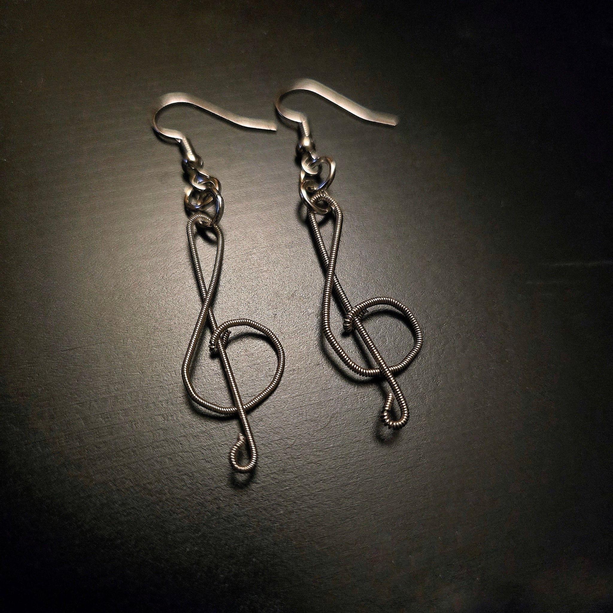 pair of earrings in the shape of treble clefs, made from upcycled guitar strings lying a black background 