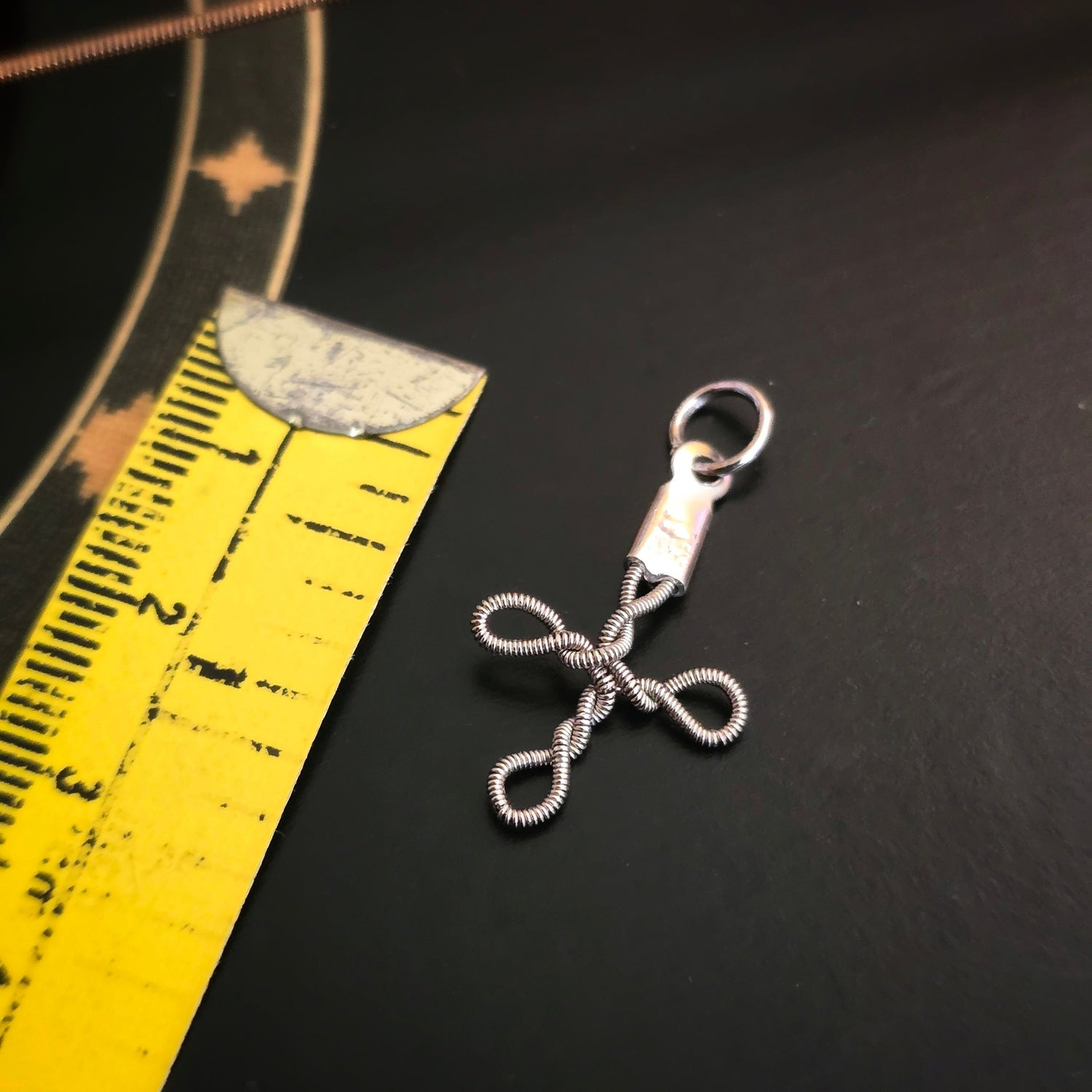 small upcycled guitar string cross style pendant on a black background with a yellow measuring tape.