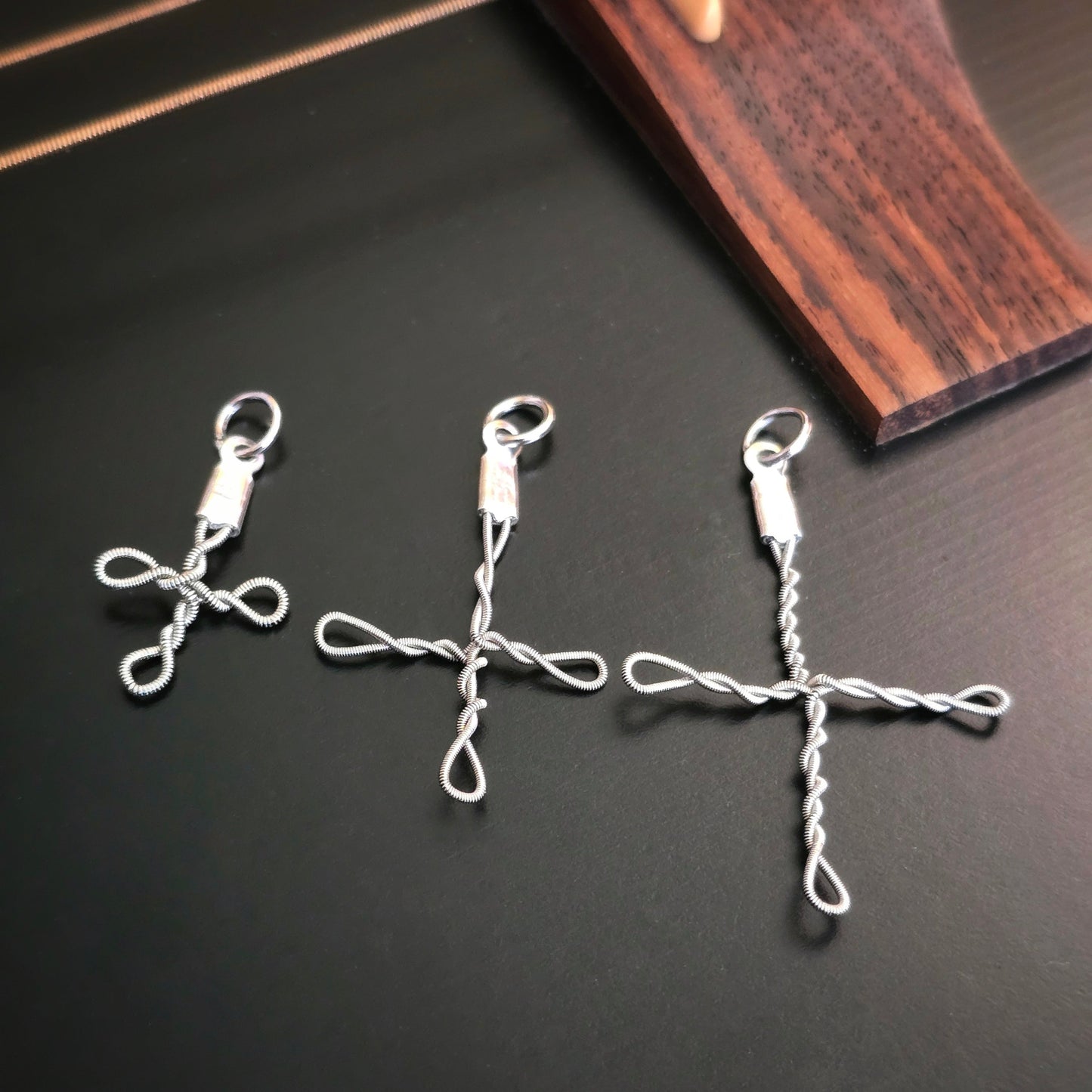Three silver upcycled guitar string cross pendants in various sizes on a dark surface with a wooden guitar neck in the background.