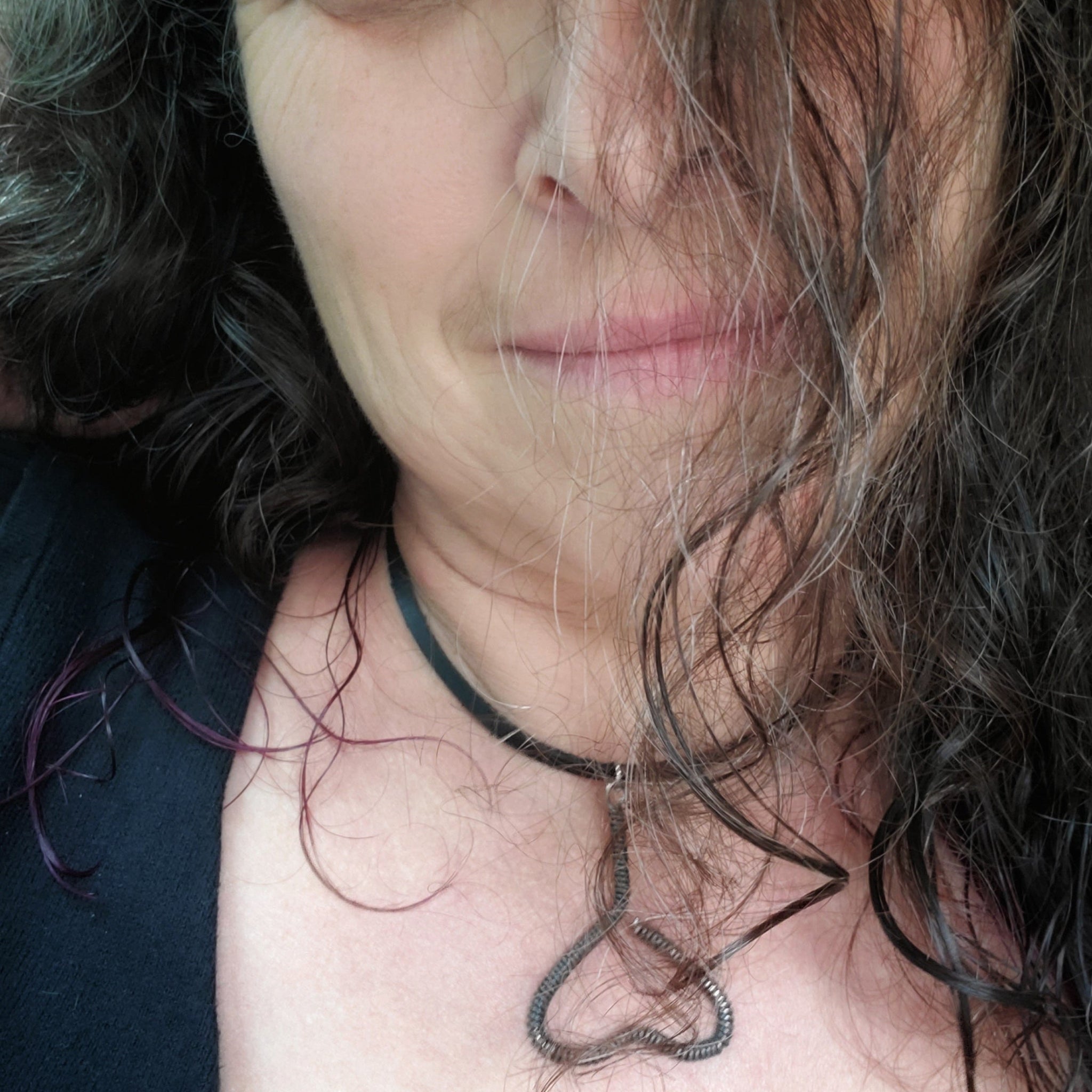 woman's head with black, grey and purple hair - she is wearing a necklace - pendant is in the shape of an upside down heart and is made from a black upcycled piano string - pendant hangs off a black leather cord