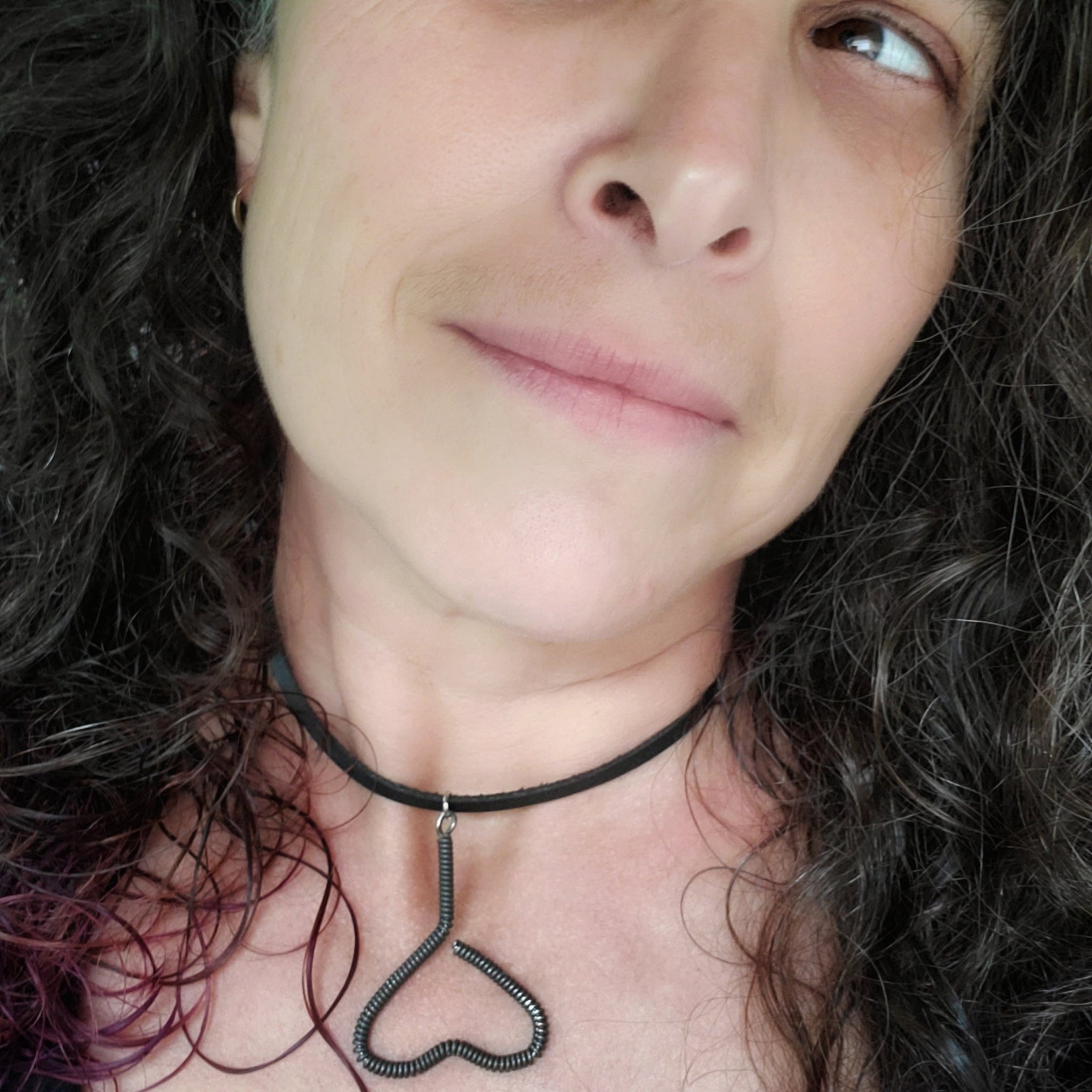 woman's head with black, grey and purple hair - she is wearing a necklace - pendant is in the shape of an upside down heart and is made from a black upcycled piano string - pendant hangs off a black leather cord