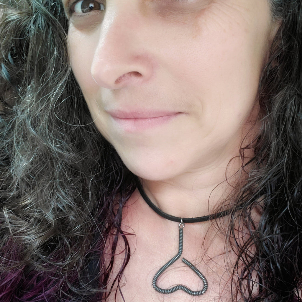 woman's head with black, grey and purple hair - she is wearing a necklace - pendant is in the shape of an upside down heart and is made from a black upcycled piano string - pendant hangs off a black leather cord