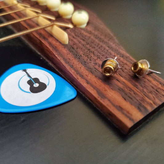 a pair of stud style earrings made from guitar string ballends sit on the bridge of a black guitar, beside the bridge is a blue guitar pick with a picture of a black guitar