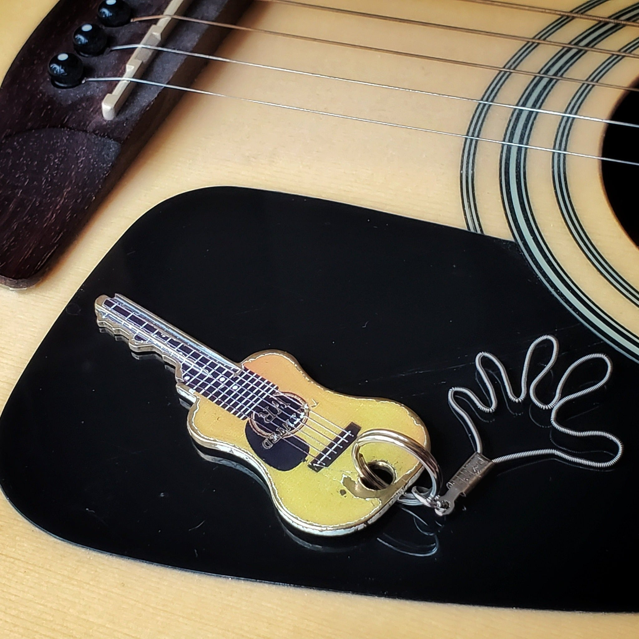 a keychain made from an upcycled guitar string shaped like a hand  tied to a key shaped like a guitar - behind it is a black pickguard of a beige guitar