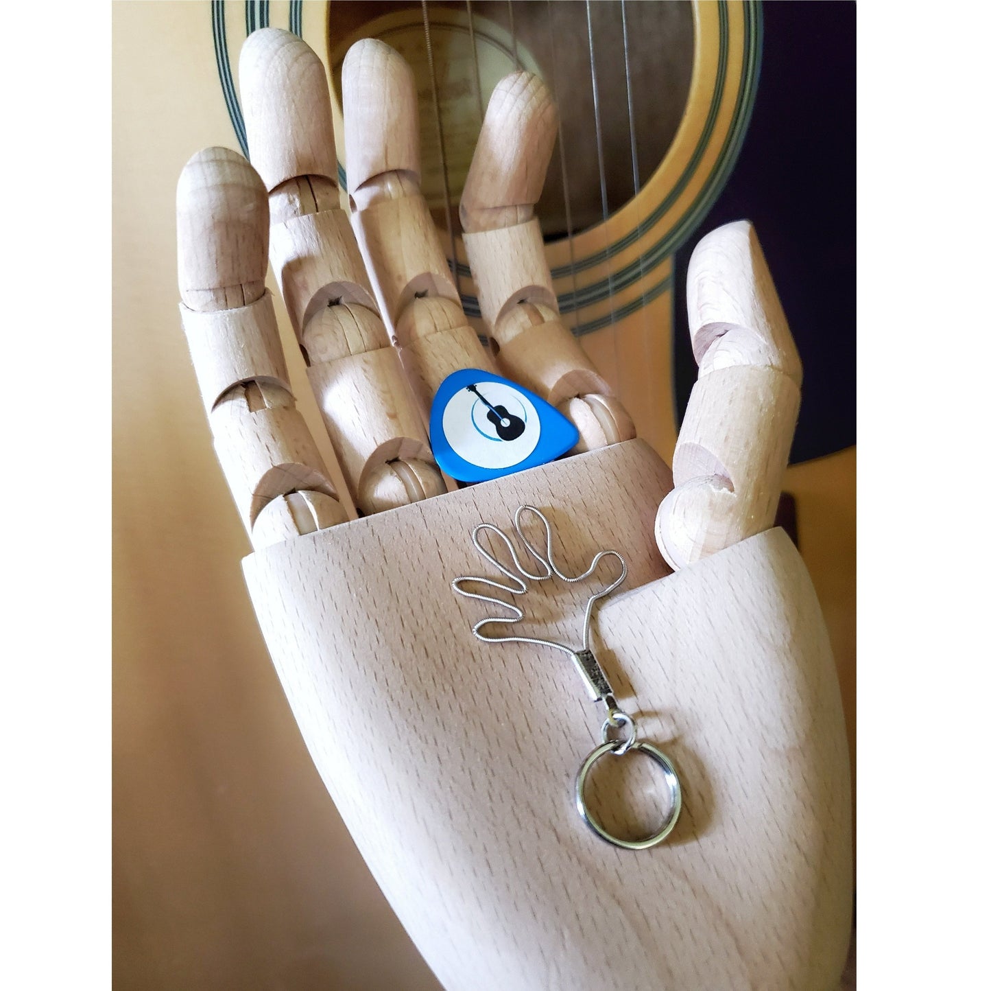 a keychain made from an upcycled guitar string shaped like a hand - above it is a blue guitar pick with an image of a black guitar - both are sitting in the palm of a wooden hand behind which is a beige guitar