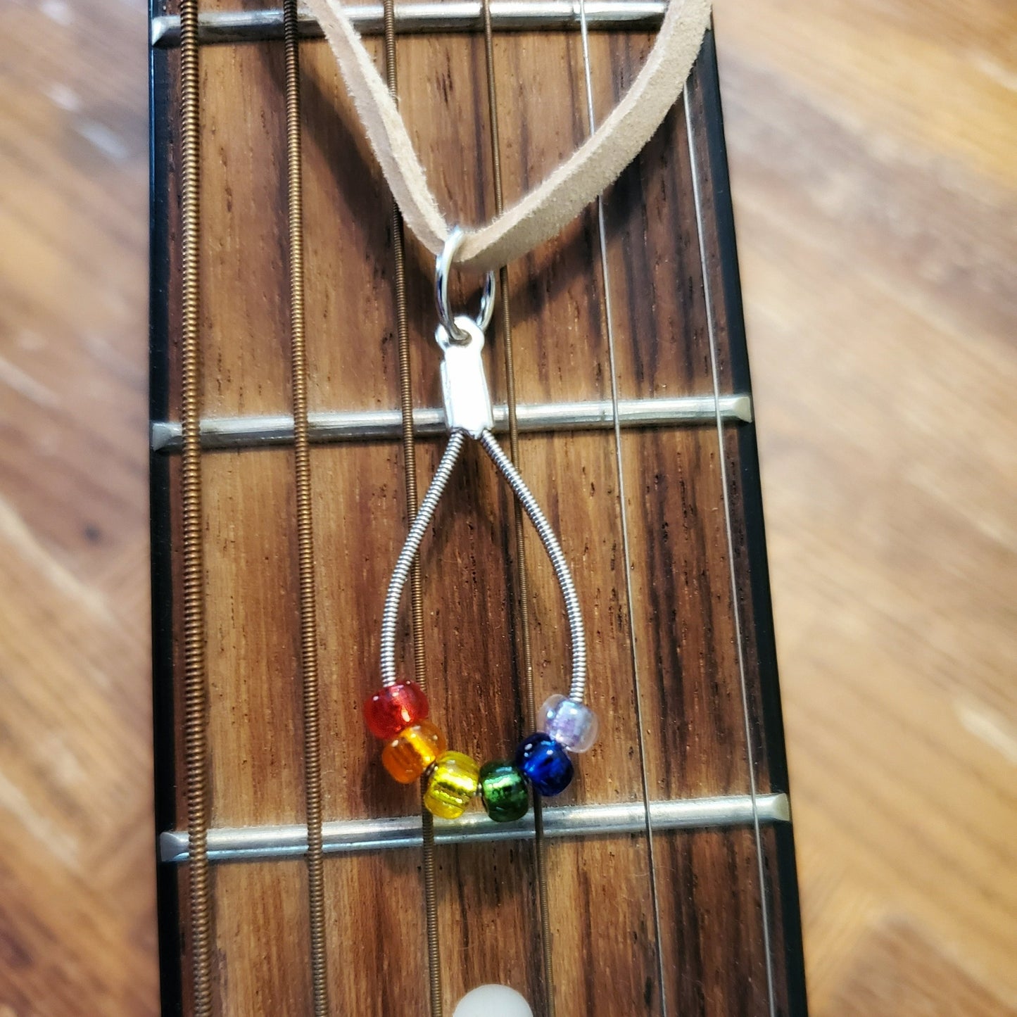 necklace made from an upcycled guitar string - 6 glass beads represent the colours of the LGBTQ flag (red, orange, yellow, green, blue and purple) and a beige suede cord on the neck of a beige guitar