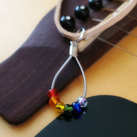 necklace made from an upcycled guitar string - 6 glass beads represent the colours of the LGBTQ flag (red, orange, yellow, green, blue and purple) and a beige suede cord on the bridge of a beige guitar