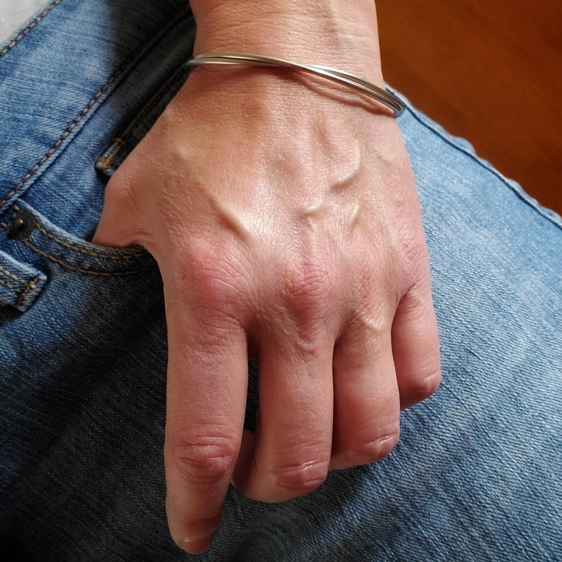 woman's hand the thumb is hook onto a jean pocket on her wrist there is a silver coloured clasp style bracelet made from upcycled upright bass strings