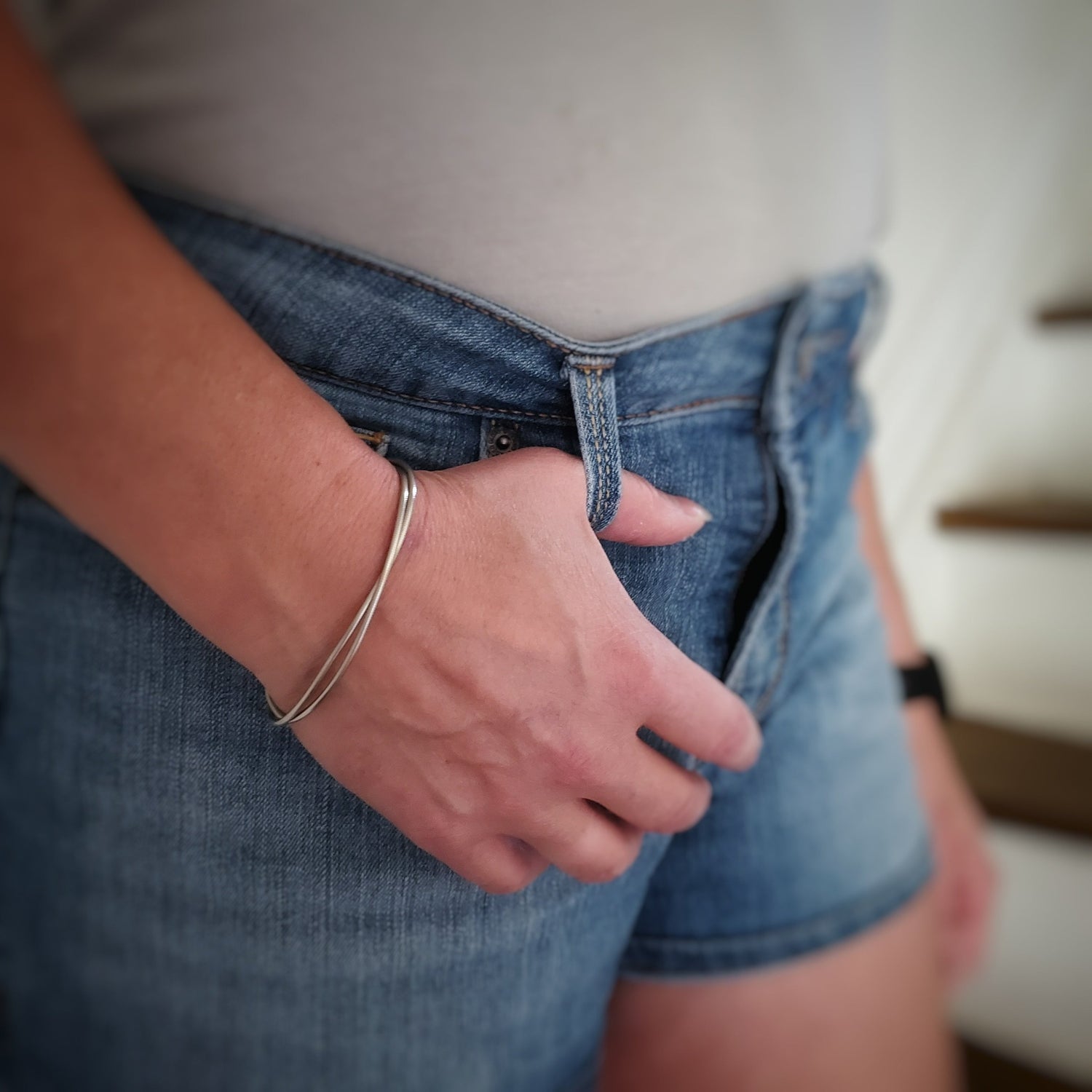 woman's arm and hand, her thumb is hooked into the loop of her jean shorts we see part of her white tshirt - on her wrist she is wearing a silver coloured clasp style bracelet made from upcycled upright bass strings