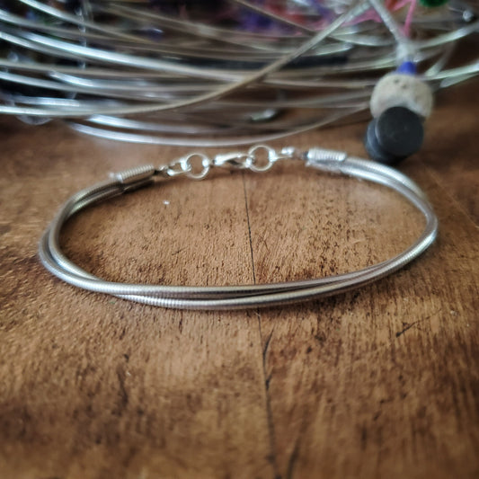 silver coloured clasp style bracelet made from upcycled upright bass strings - behind is a pile of upright bass strings