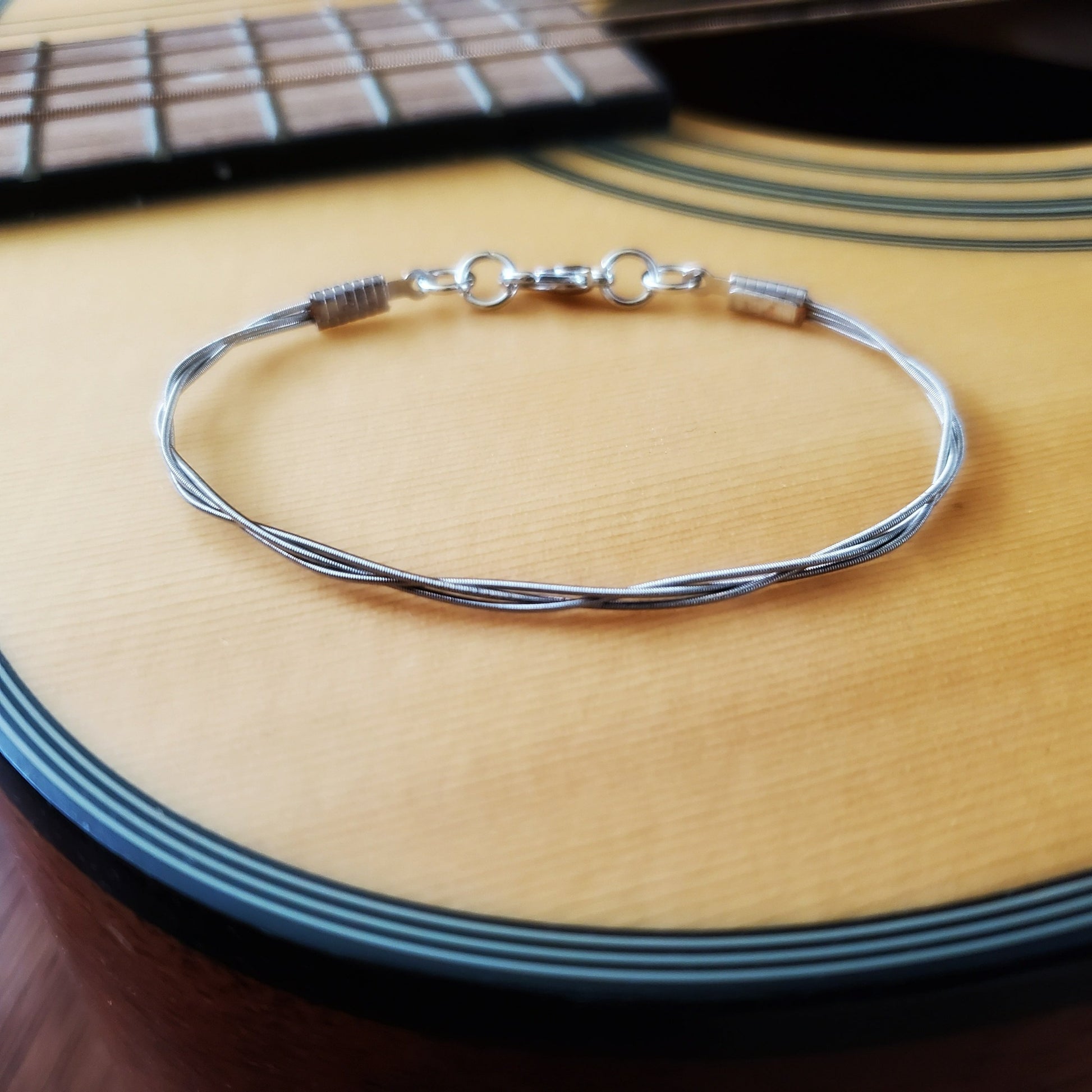 bracelet made from 3 strands of guitar strings braided together sitting on the body of a tan coloured acoustic guitar