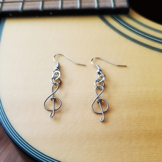 pair of earrings in the shape of treble clefs, made from upcycled guitar strings lying on the body of a beige guitar