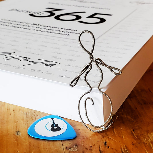 bookmark in the shape of a woman's form - made from an upcycled guitar string - it is resting on the corner of a white book with black words on it- next to it is a blue guitar pick with an image of a black guitar