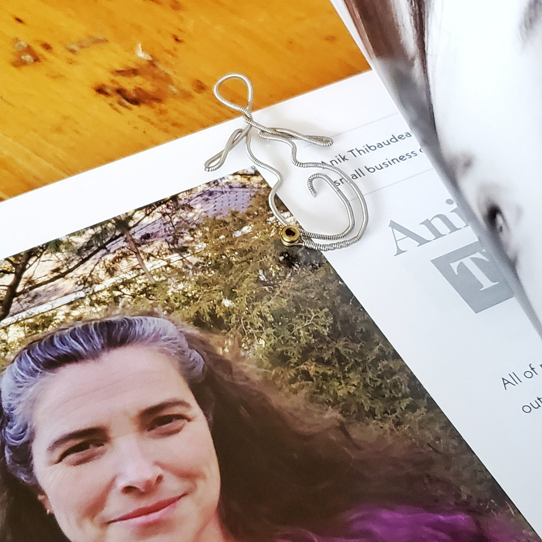 bookmark in the shape of a woman's form - made from an upcycled guitar string - it is resting on a page from a book on which we can see the face of a woman with long grey, brown and black hair and some black or grey words on a white page