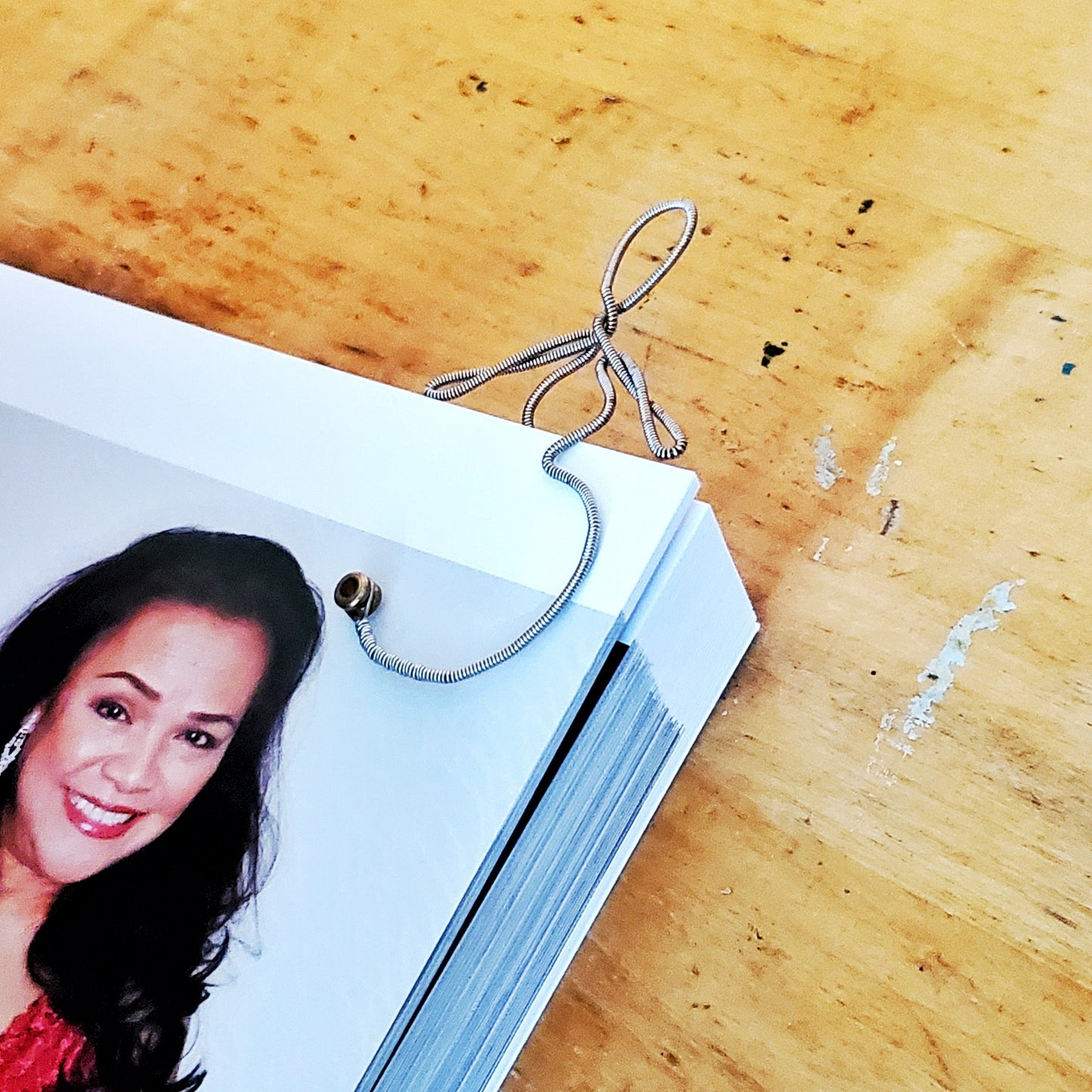 bookmark in the shape of a woman's form - made from an upcycled guitar string it is hooked onto the corner of a page from a book on which we can see the face of a woman with long dark hair who is smiling
