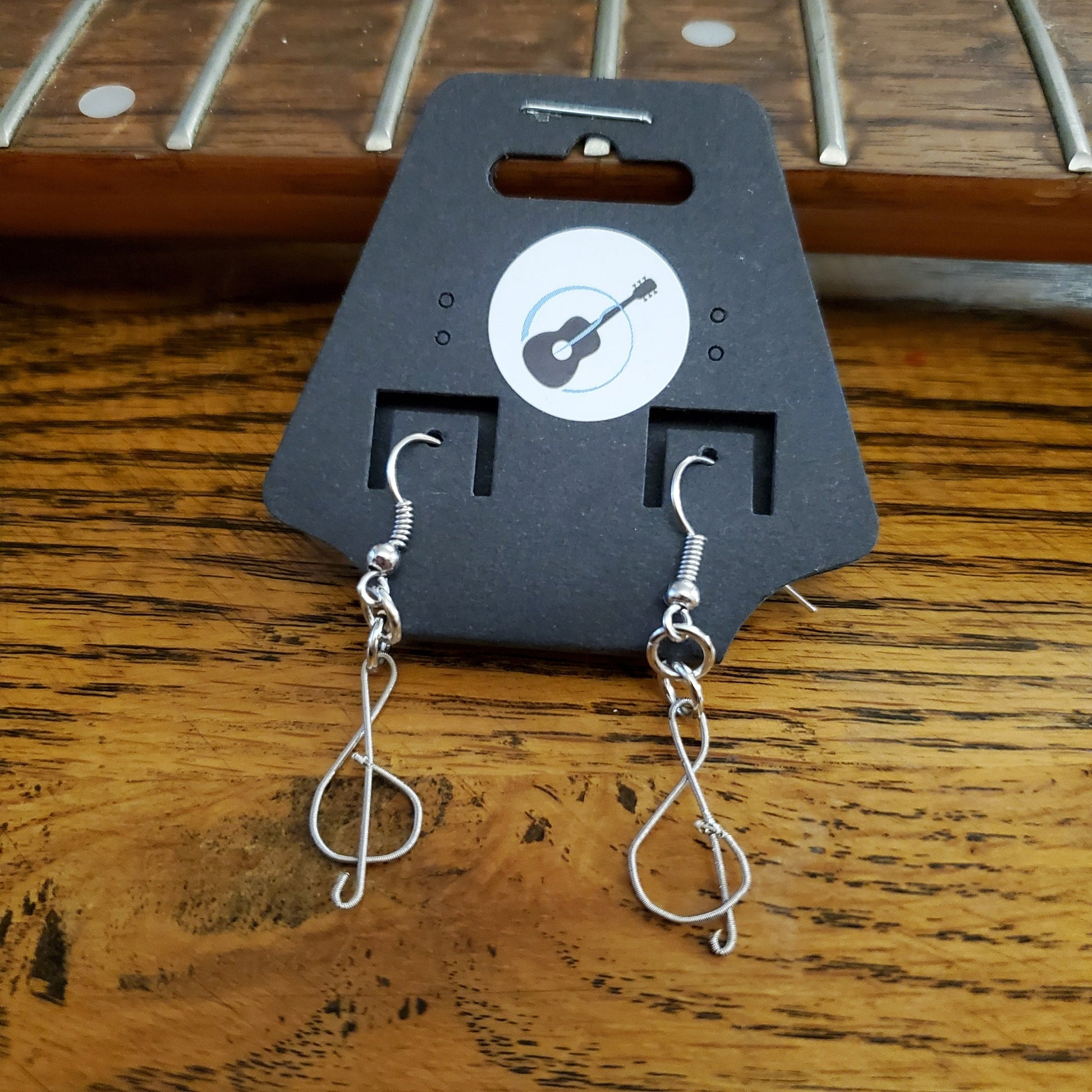 pair of earrings in the shape of treble clefs, made from upcycled guitar strings, hanging from a black earring card lying on the neck of a guitar which as no strings