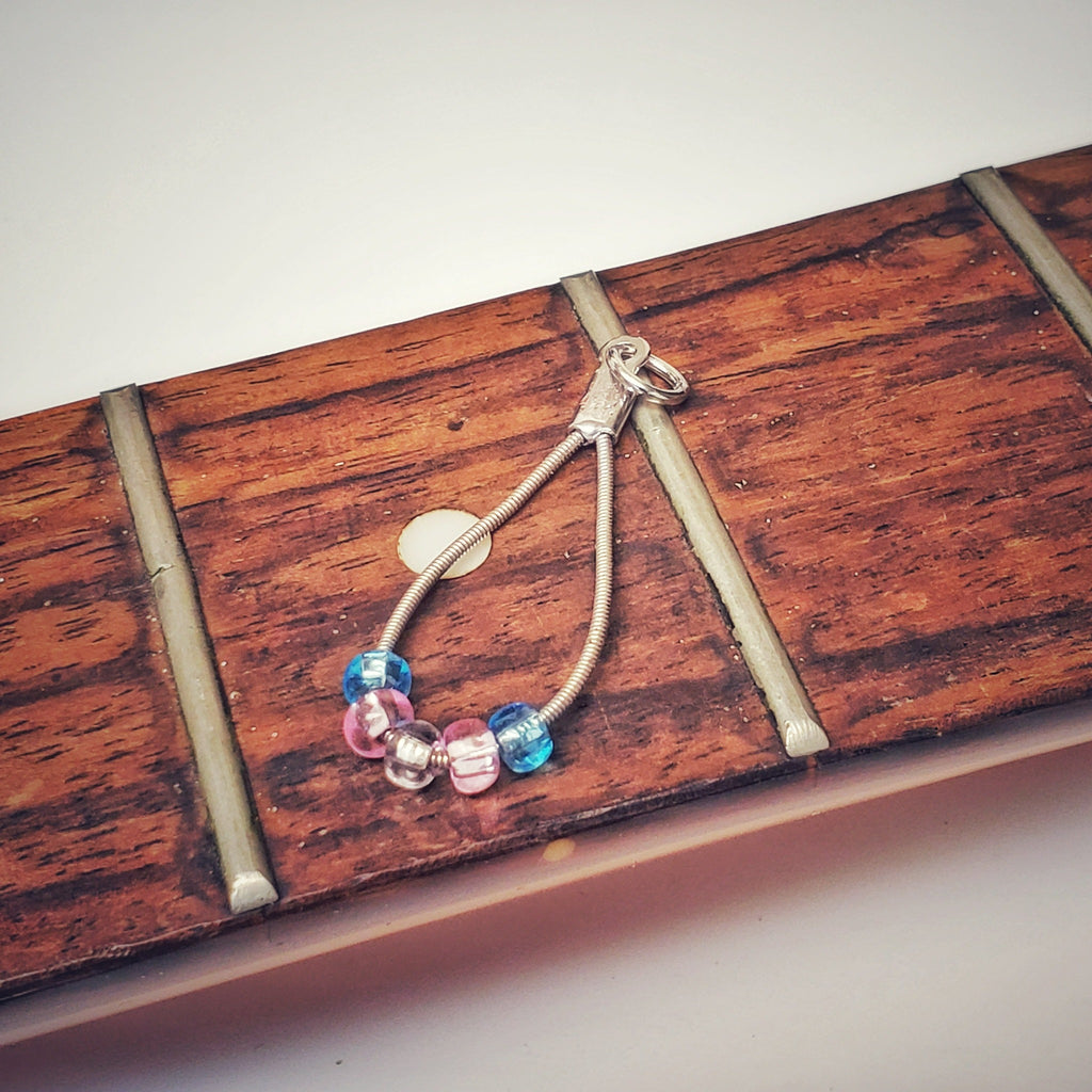 silver coloured teardrop pendant made from upcycled guitar strings - there are 5 beads representing the colours of the transgender pride flag - 2 blue, 2 pink and 1 white - pendant is sitting on the neck of a guitar which has no stings