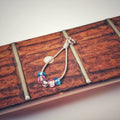 silver coloured teardrop pendant made from upcycled guitar strings - there are 5 beads representing the colours of the transgender pride flag - 2 blue, 2 pink and 1 white - pendant is sitting on the neck of a guitar which has no stings