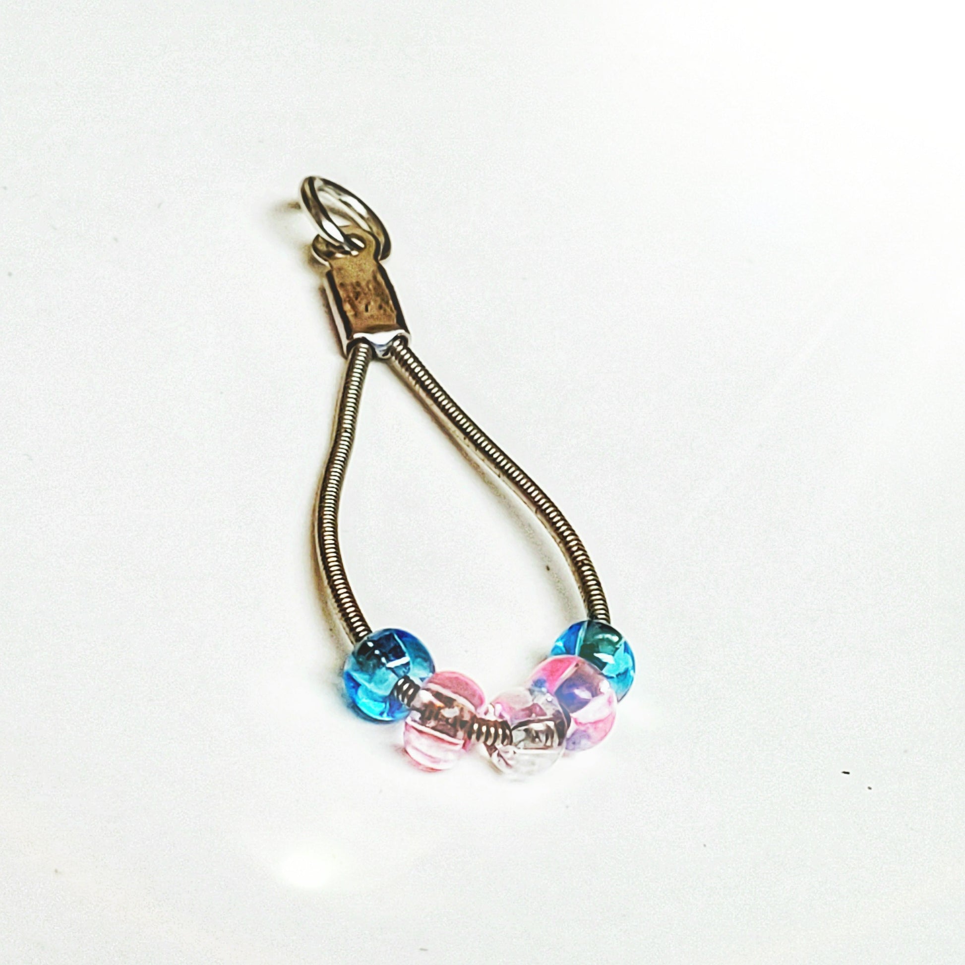 silver coloured teardrop pendant made from upcycled guitar strings - there are 5 beads representing the colours of the transgender pride flag - 2 blue, 2 pink and 1 white - white background