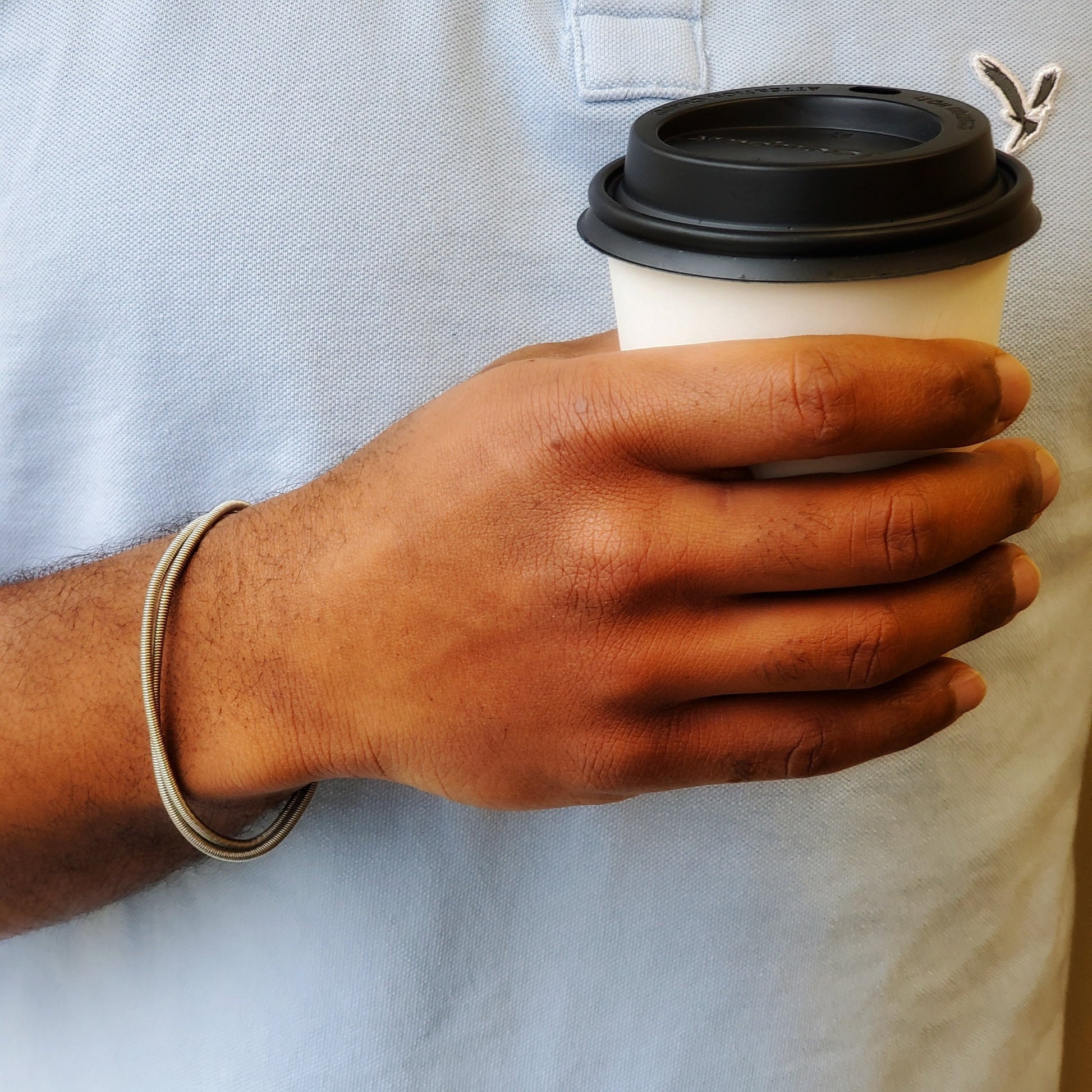 man's hand and wrist - he is holding a black and white coffee cup - on his wrist he is wearing a silver coloured bracelet made from 2 strands of upcycled electric bass strings