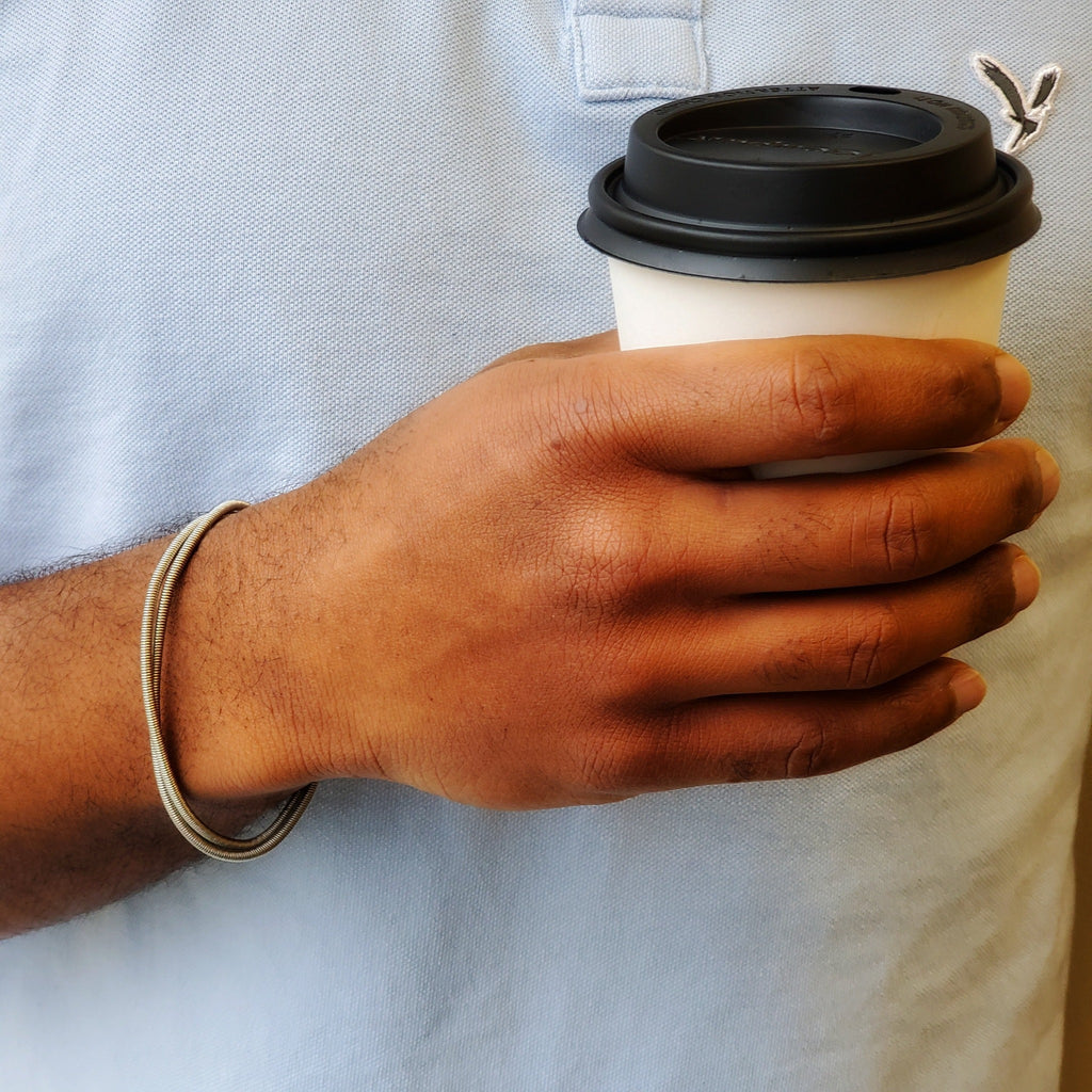 man's hand and wrist - he is holding a black and white coffee cup - on his wrist he is wearing a silver coloured bracelet made from 2 strands of upcycled electric bass strings