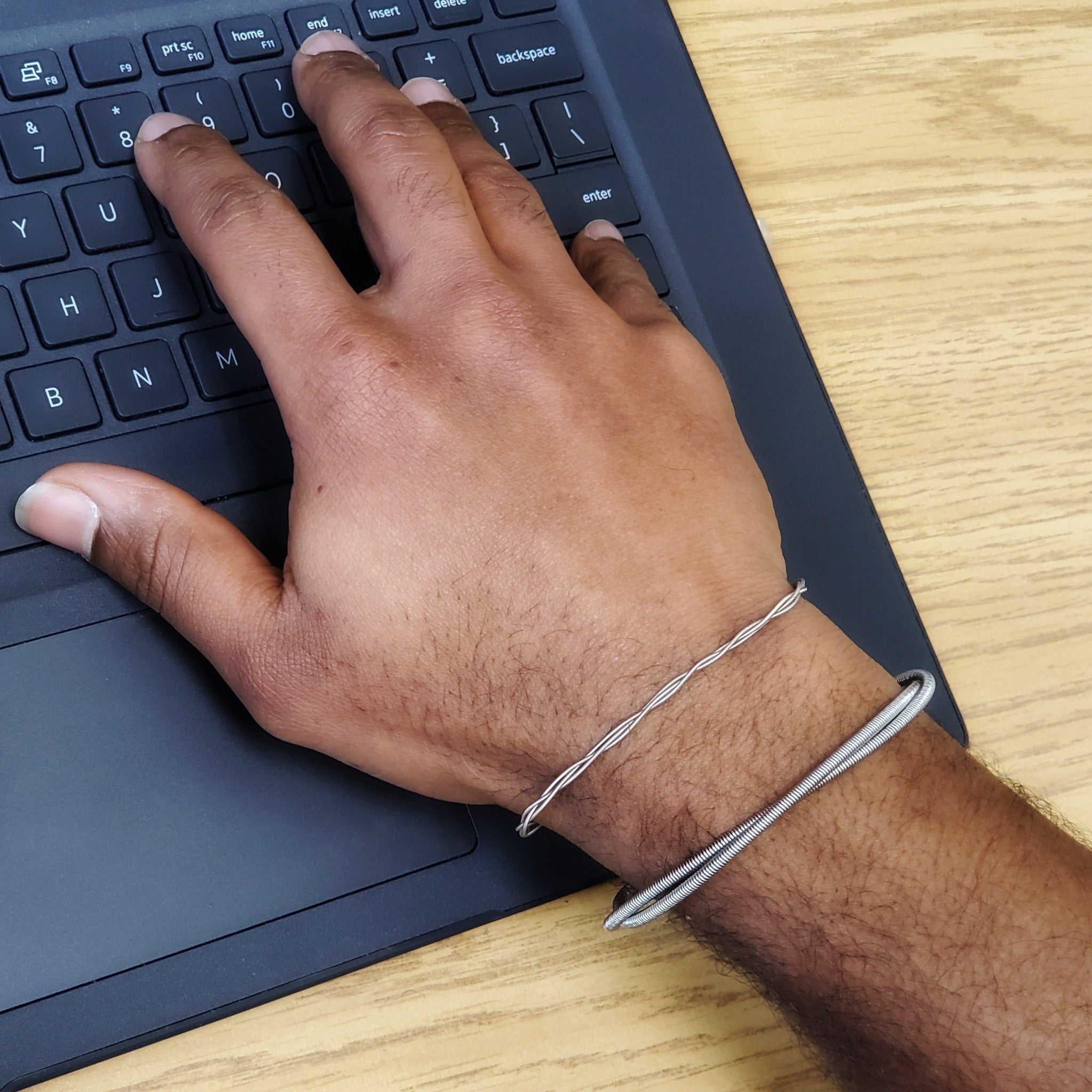 man's hand and wrist on his wrist he is wearing a silver coloured bracelet made from 2 strands of upcycled electric bass strings as well as a thinner bracelet made from braided guitar strings - his fingers are resting on a computer keyboard