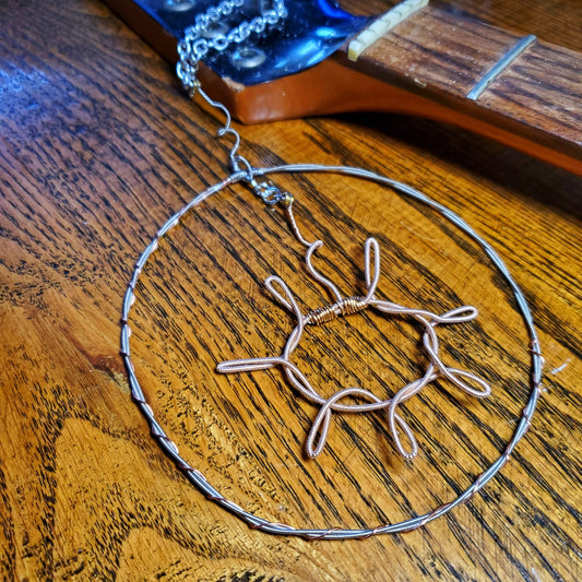 a decoration shaped like a sun with a circle around it hanging off a chain - the sun and circle are made from upcycled guitar strings - above there is a guitar neck with no strings - wood grain background