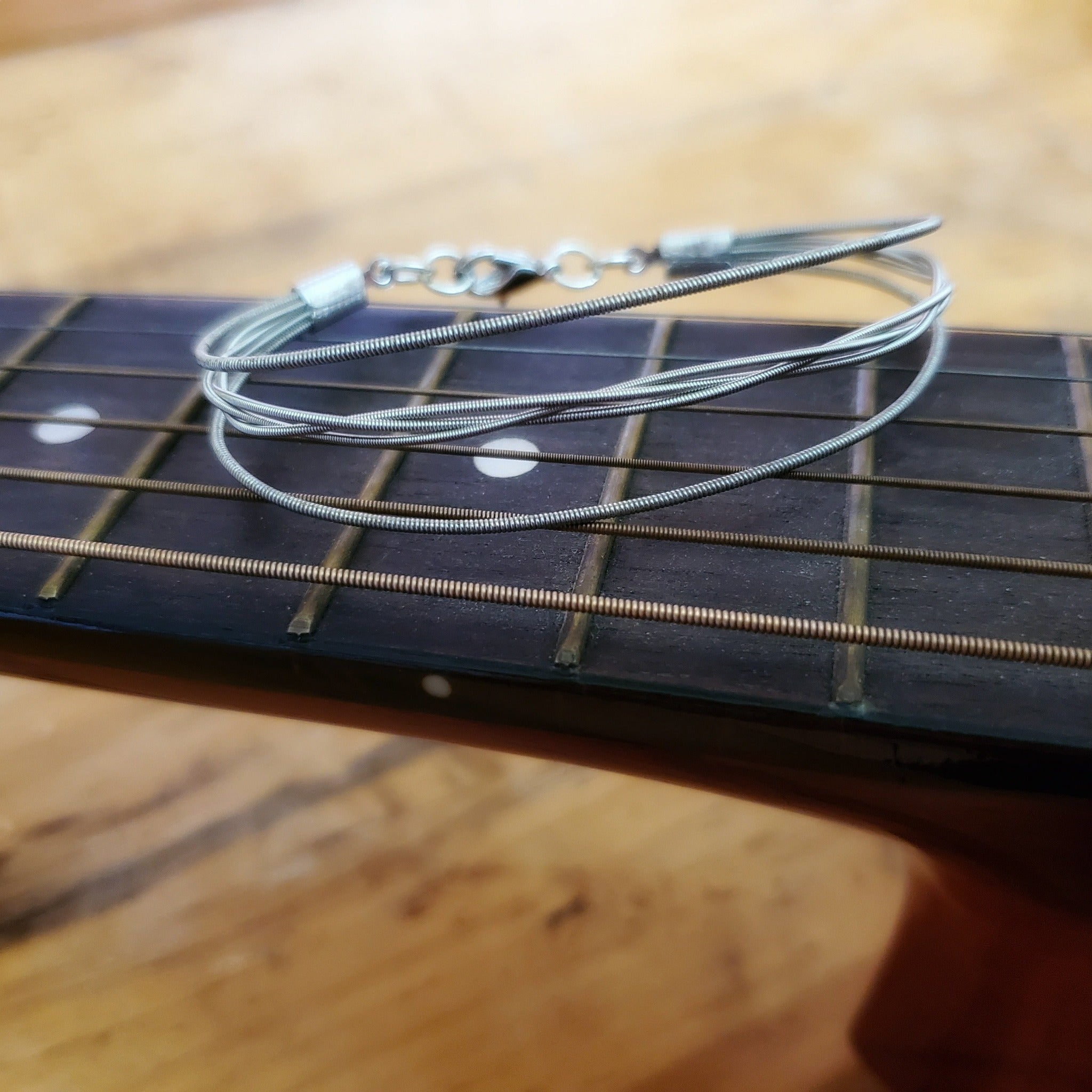 a silver coloured bracelet - between 2 guitar strings, 3 others are braided - the bracelet rests on the neck of a guitar