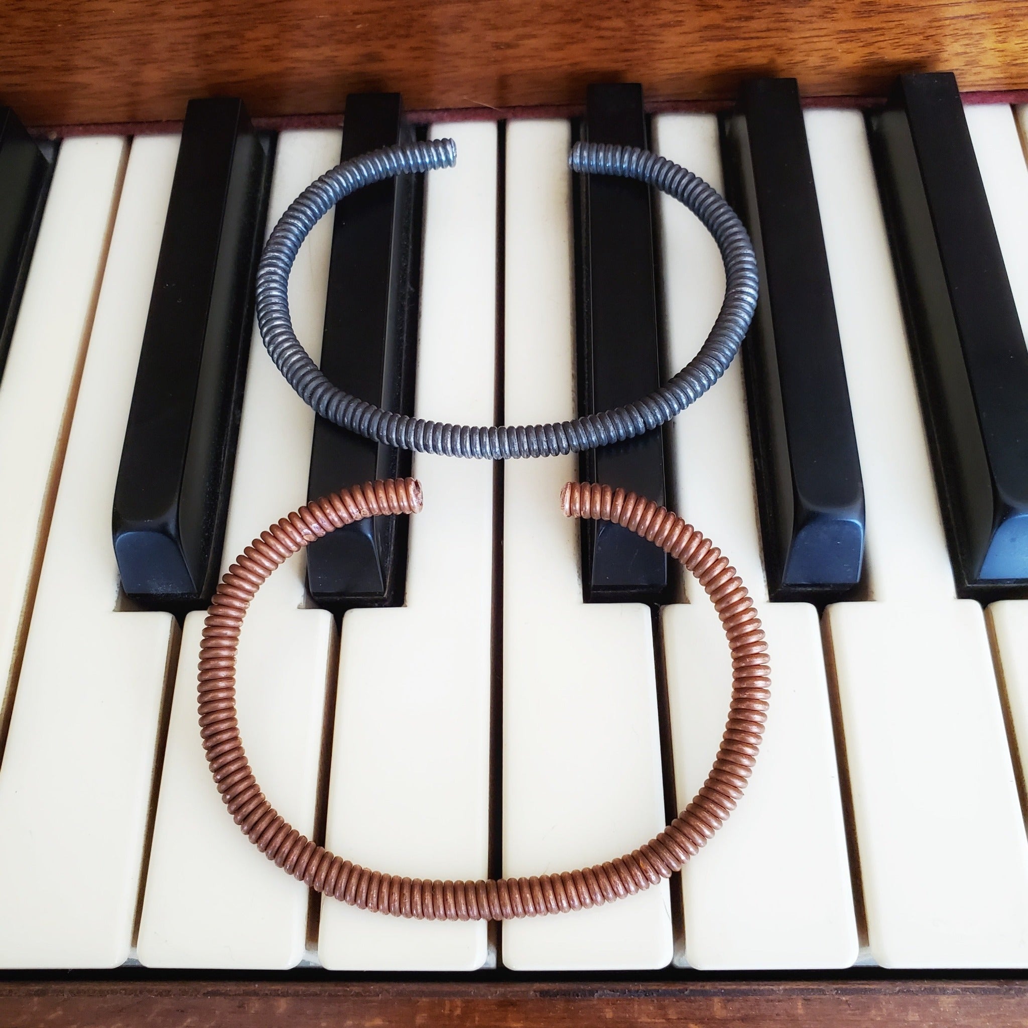copper and charcoal grey cuff style bracelets made from an upcycled piano strings - both bracelets are lying on piano keys