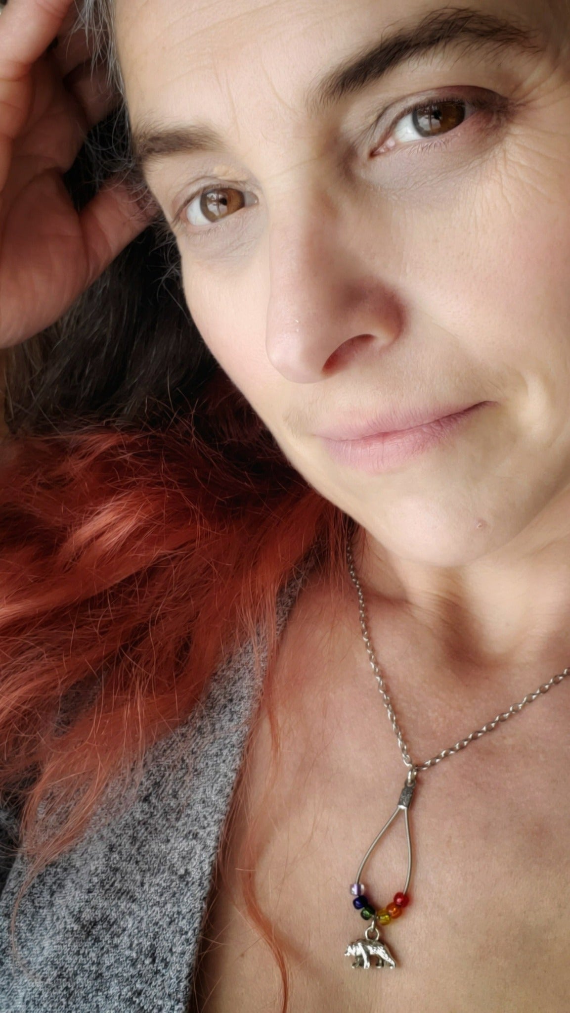 face of a woman with red and brown hair wearing a chain style necklace with a teardrop shaped pendant made from an upcycled guitar string and six glass beads representing the colours of the LGBTQ pride flag and a bear shaped silver coloured pendant
