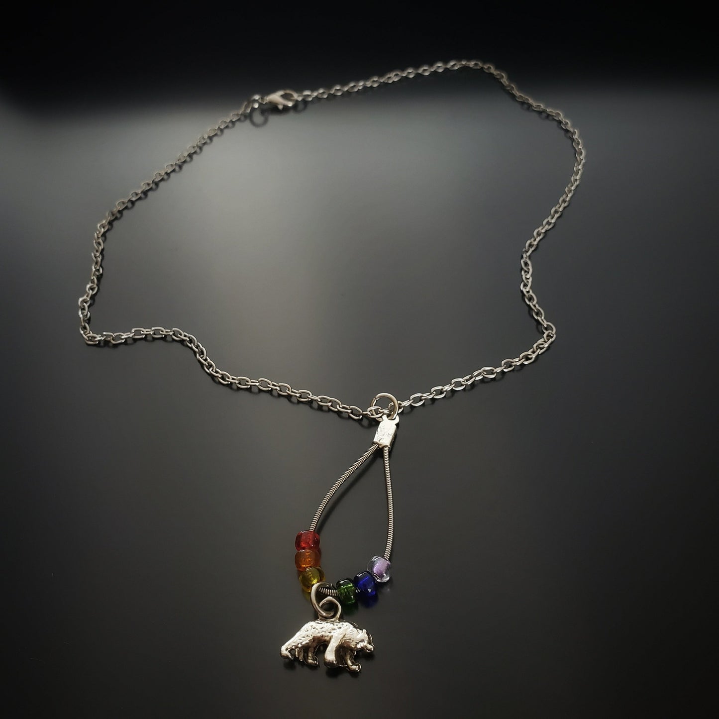 chain style necklace with a teardrop shaped pendant made from an upcycled guitar string and six glass beads representing the colours of the LGBTQ pride flag and a bear shaped silver coloured pendant