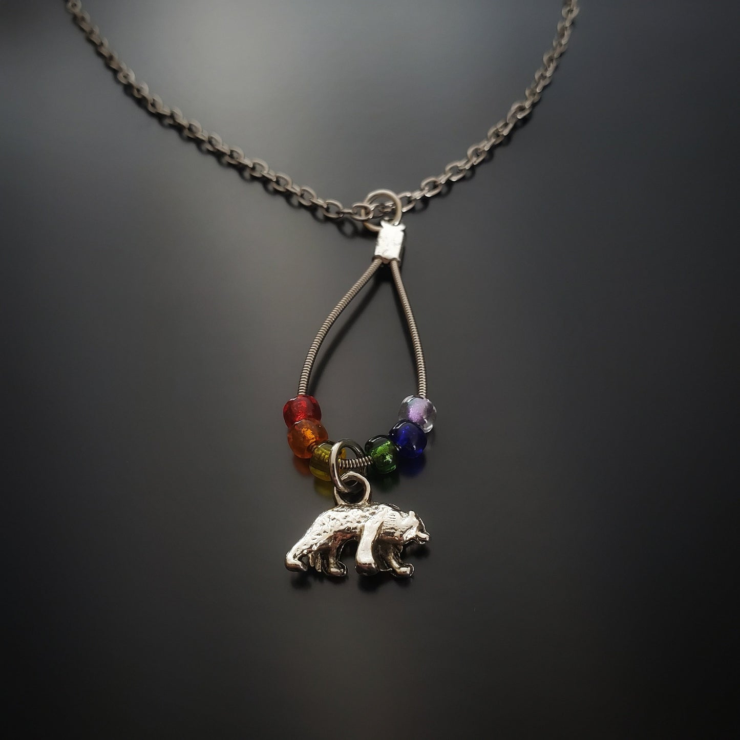 close-up of a chain style necklace with a teardrop shaped pendant made from an upcycled guitar string and six glass beads representing the colours of the LGBTQ pride flag and a bear shaped silver coloured pendant