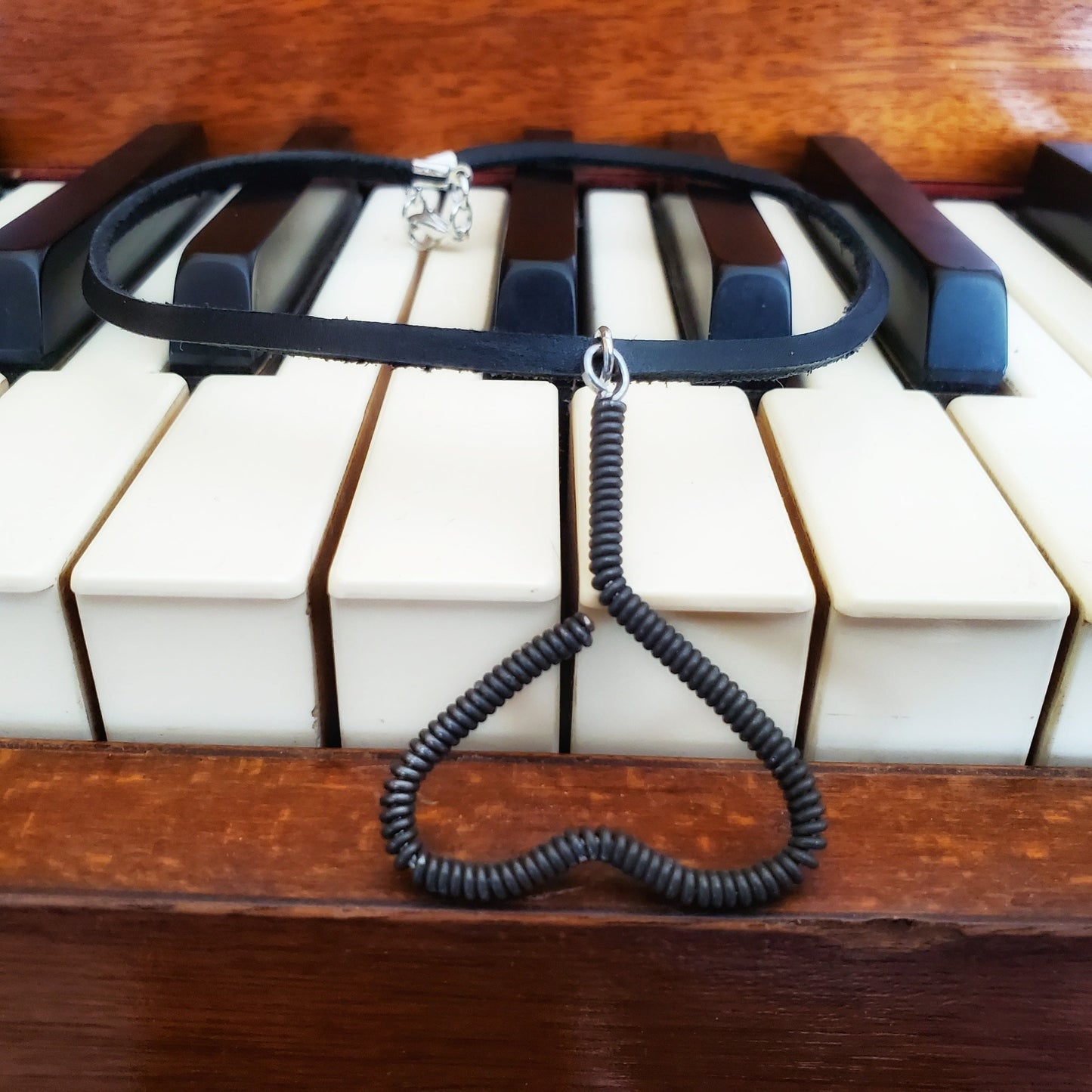 necklace - pendant is in the shape of an upside down heart and is made from a black upcycled piano string - pendant hangs off a black leather cord- necklace is lying on a piano keyboard