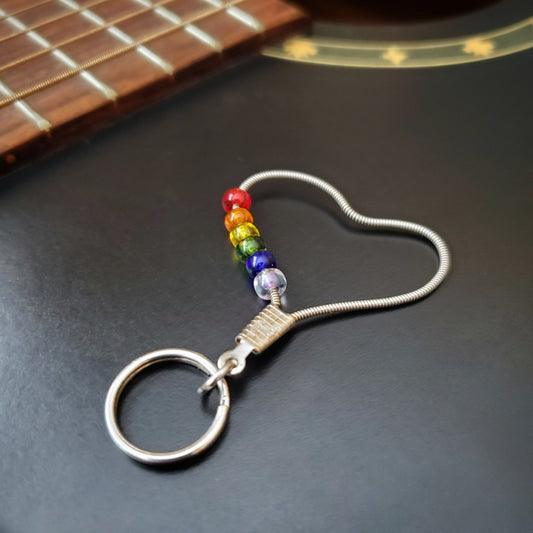 keychain made from an upcycled guitar string - 6 glass beads represent the colours of the LGBTQ flag (red, orange, yellow, green, blue and purple) - on a black guitar body